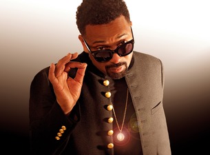 Man in dark sunglasses and a black coat with brass buttons adjusts his glasses, looking at the camera.
