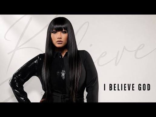 Model with long black hair wearing a black outfit, posing against a light background with the phrase 'I BELIEVE GOD' to the right.