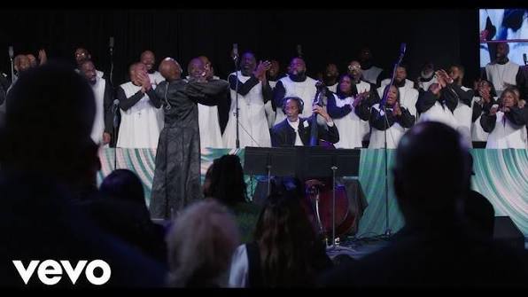 Choir in white robes singing on stage with conductor in front, audience silhouettes in foreground; Vevo watermark bottom-left.