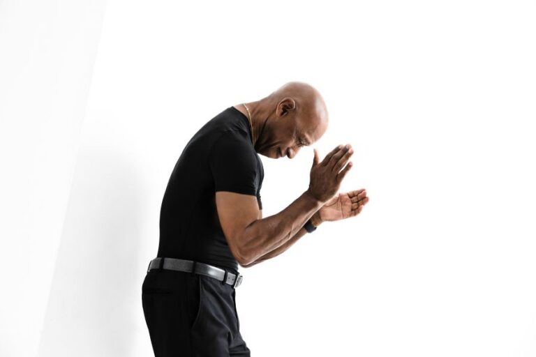 Bald man in black athletic wear bows with palms pressed together in front of him on a white background.